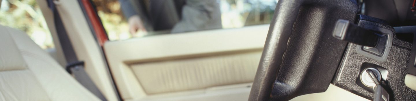 A close-up view of a car interior showing a key fob held in a person's hand near the steering wheel, with the driver's seat and door visible. Location: Winston-Salem, NC.