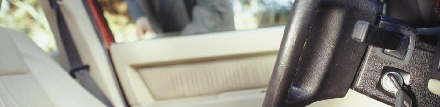 A close-up view of a car interior showing a key fob held in a person's hand near the steering wheel, with the driver's seat and door visible. Location: Winston-Salem, NC.